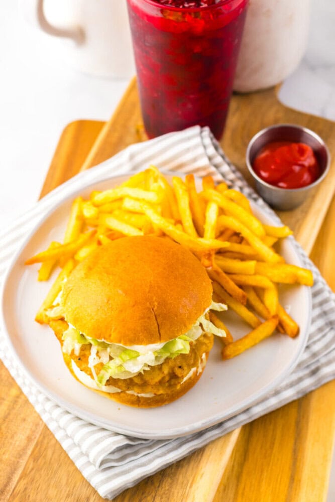 A chicken sandwich with shredded lettuce, a side of French fries, a small cup of ketchup, and a red beverage on a wooden board.