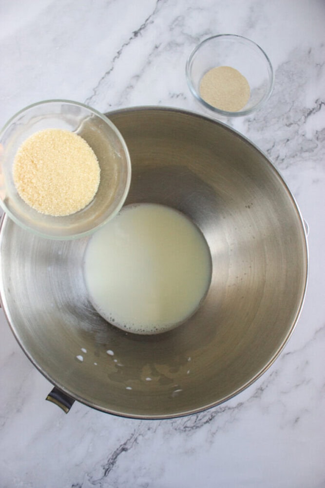 A bowl of milk in a metal mixing bowl, with a small glass bowl of sugar being poured in, and another small bowl containing yeast nearby.