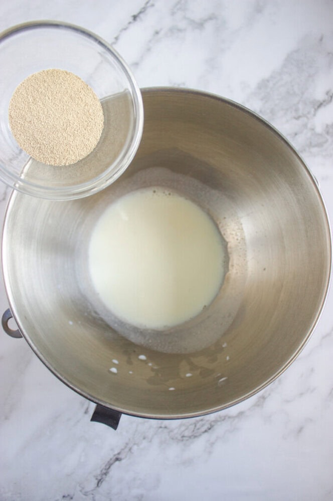 A metal mixing bowl with milk inside and a small glass bowl of dry yeast being held above it, on a marble surface.