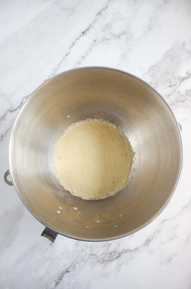 A stainless steel mixing bowl containing a partially mixed light batter sits on a white marble surface.