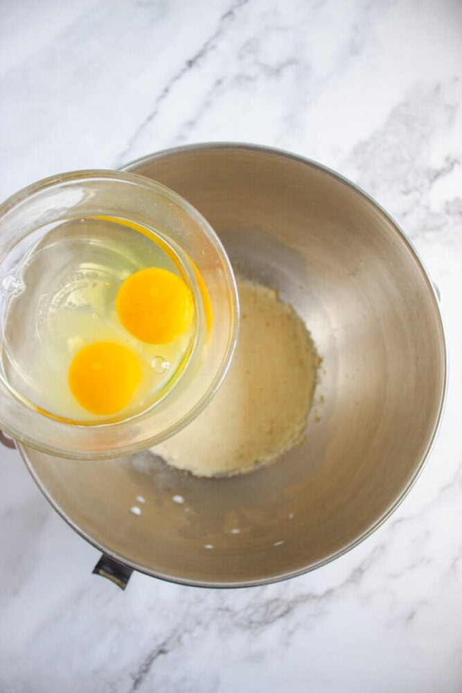 A glass measuring cup with two raw eggs is held above a metal mixing bowl containing a beige mixture on a marble countertop.