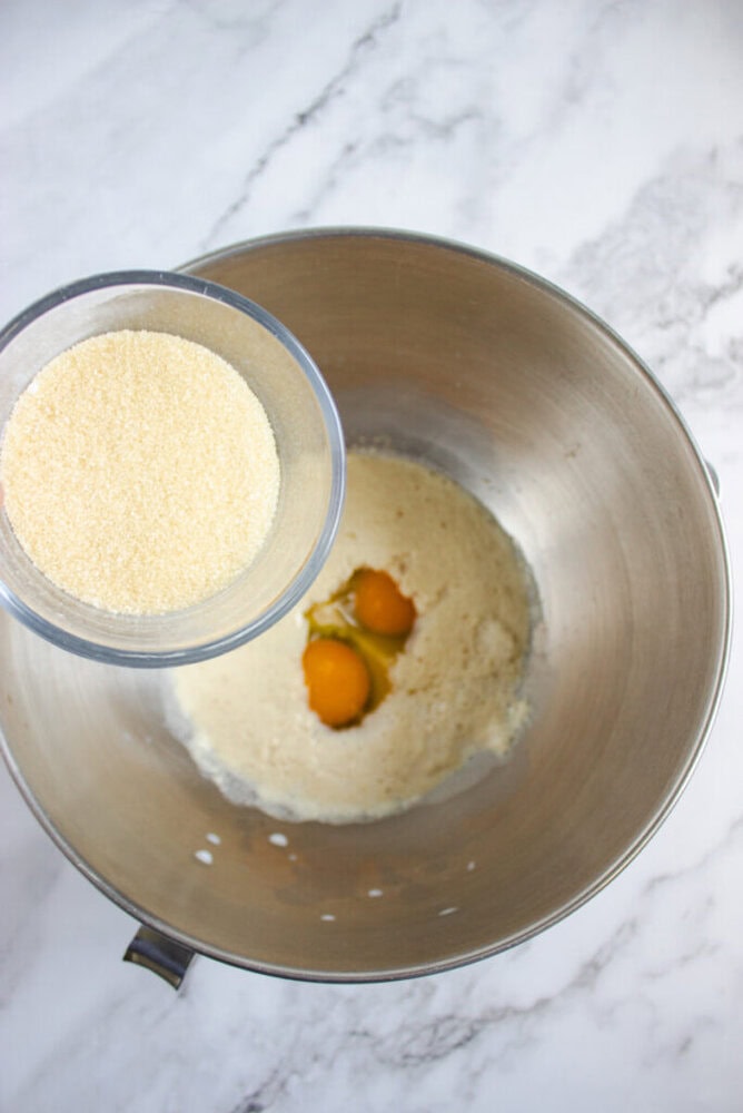 A metal mixing bowl with eggs and sugar inside, and a smaller bowl of granulated sugar being held above it, on a marble countertop.