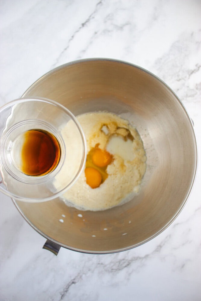 A metal mixing bowl with sugar, eggs, and milk inside, while a hand holds a cup of vanilla extract above the bowl, ready to pour it in.