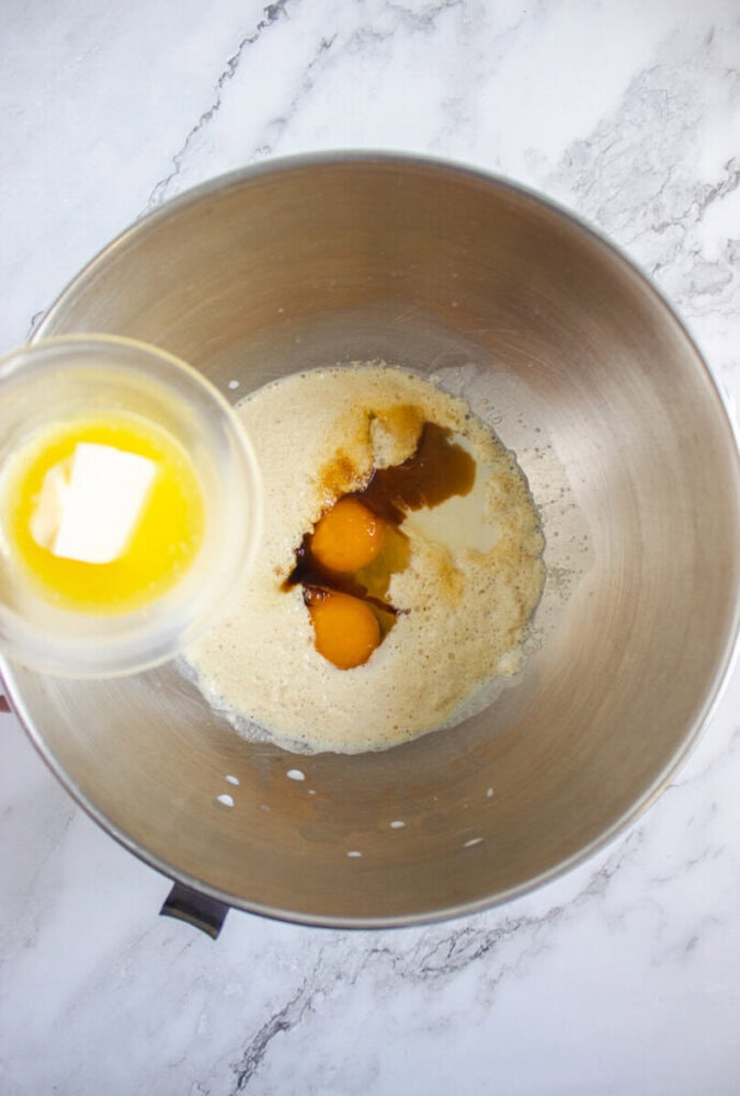 A metal mixing bowl contains eggs, sugar, and vanilla. A hand holds a measuring cup of melted butter above, about to pour it in.