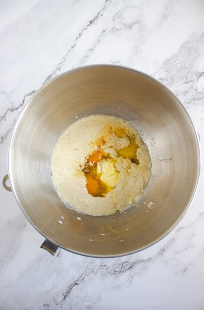 A mixing bowl contains sugar, eggs, butter, and vanilla extract on a white marble surface, ready to be mixed.