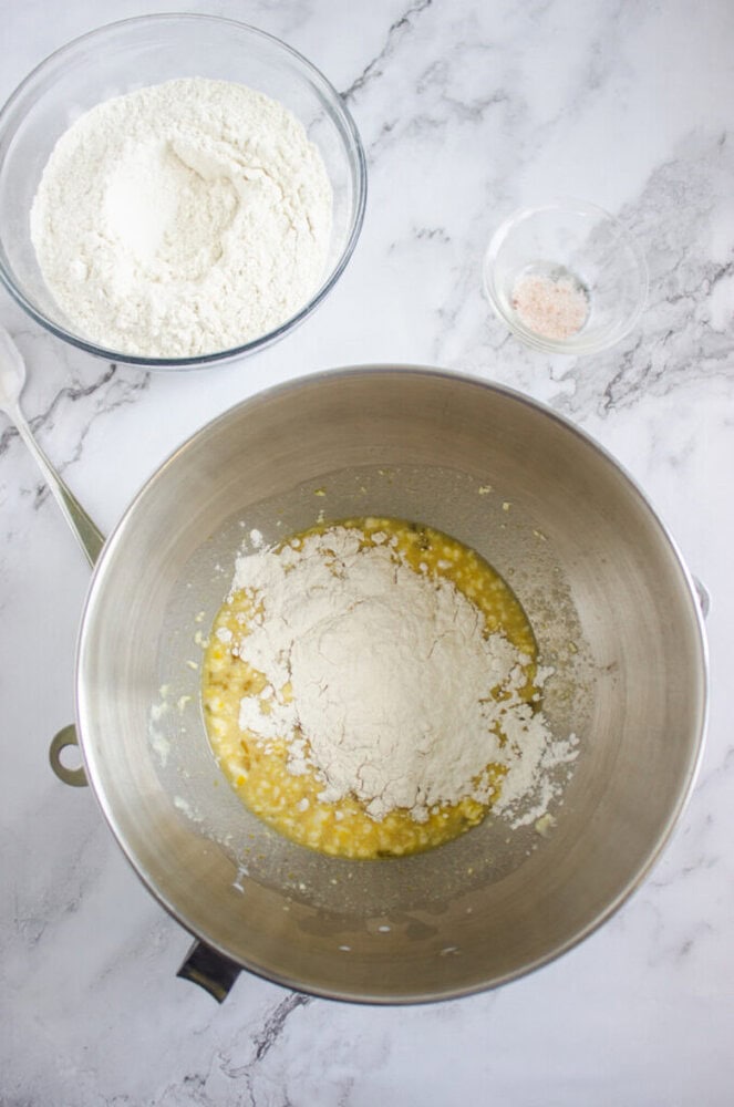 A metal mixing bowl contains mashed bananas, flour, and wet ingredients. Nearby are a glass bowl of flour and a small glass bowl of salt on a marble countertop.