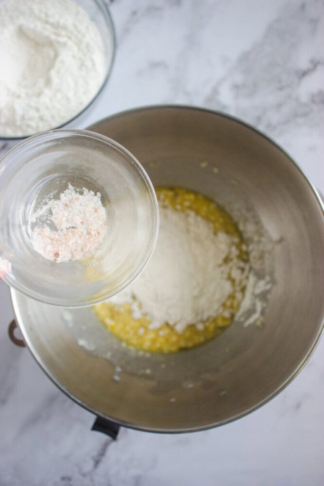A glass bowl with pink salt is held above a mixing bowl containing flour and a yellow batter mixture, with another bowl of flour in the background.