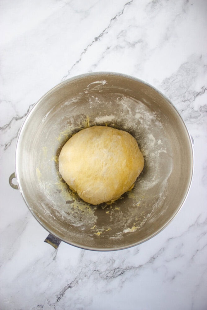 A ball of dough rests in the center of a metal mixing bowl on a white marble countertop.