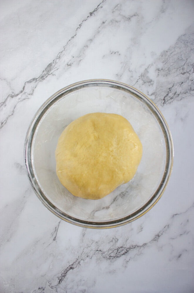Ball of yellow dough resting in a clear glass bowl on a white and gray marble surface.