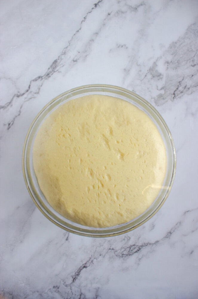 A glass bowl filled with risen dough sits on a white marble surface.