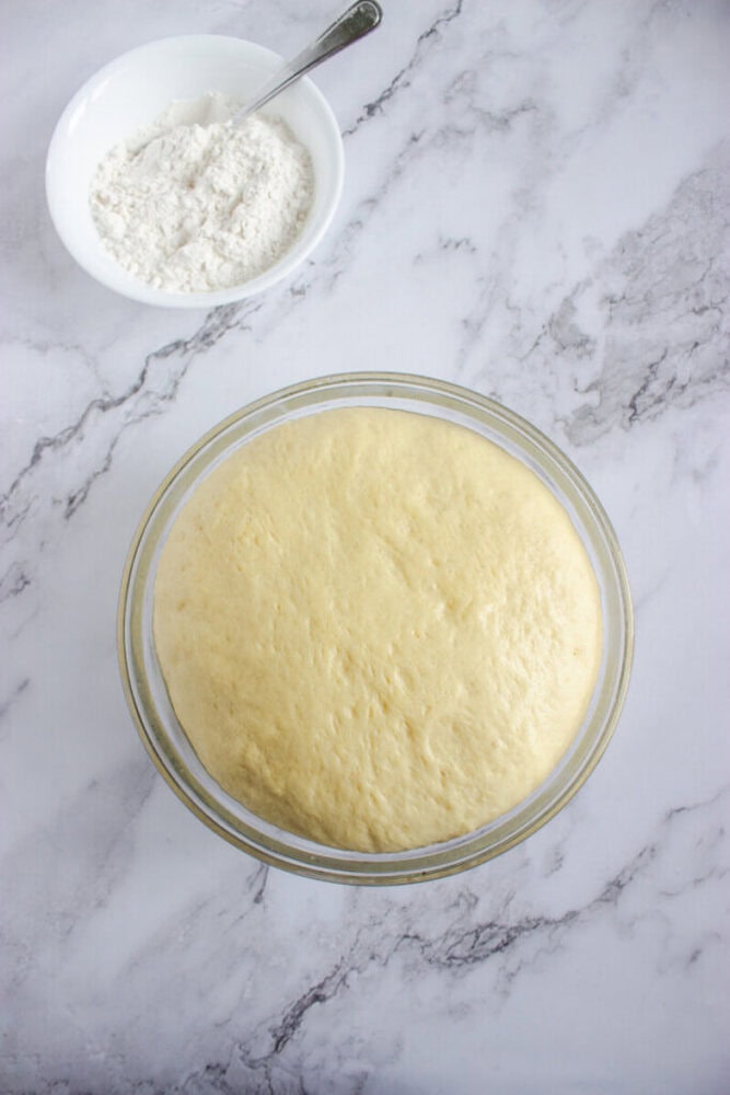 A glass bowl of risen dough sits on a marble surface next to a white bowl of flour with a metal spoon.