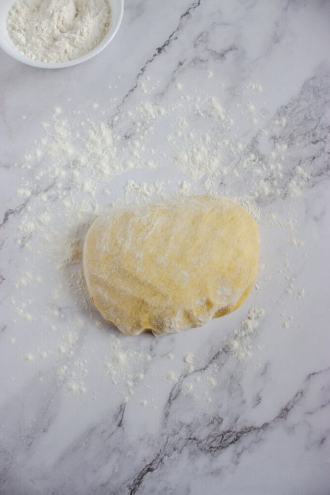 A ball of dough dusted with flour rests on a floured marble surface, with a bowl of flour visible in the upper left corner.