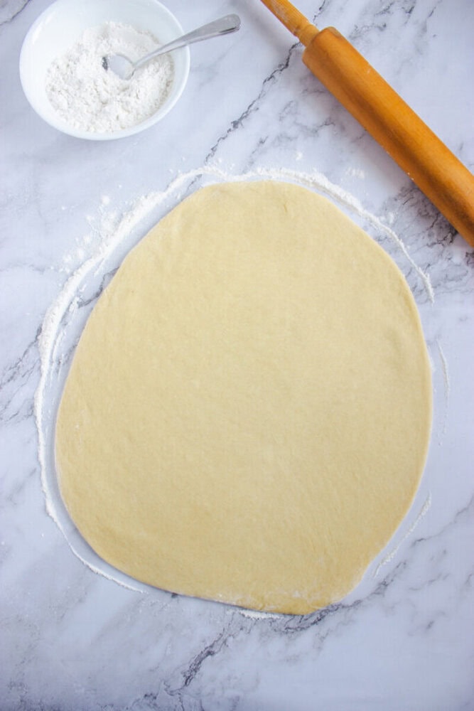 Rolled out dough on a floured marble surface with a rolling pin and a bowl of flour nearby.