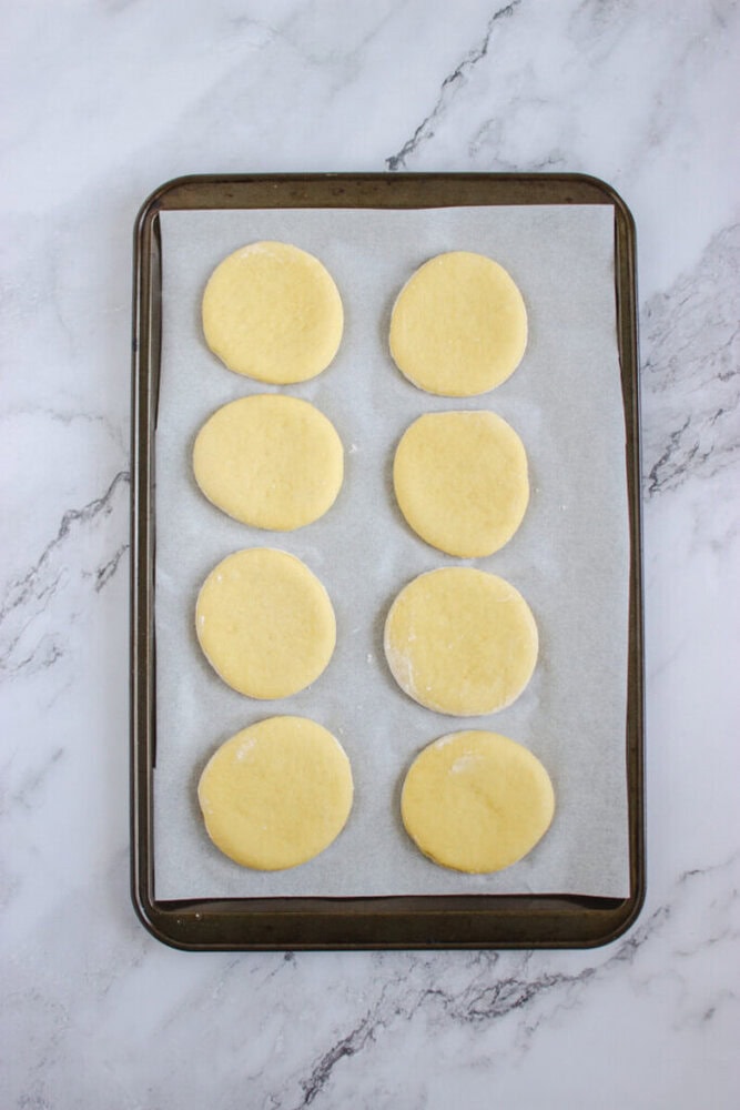 Eight round, unbaked cookie dough pieces are arranged in two columns on a parchment-lined baking sheet atop a marble surface.