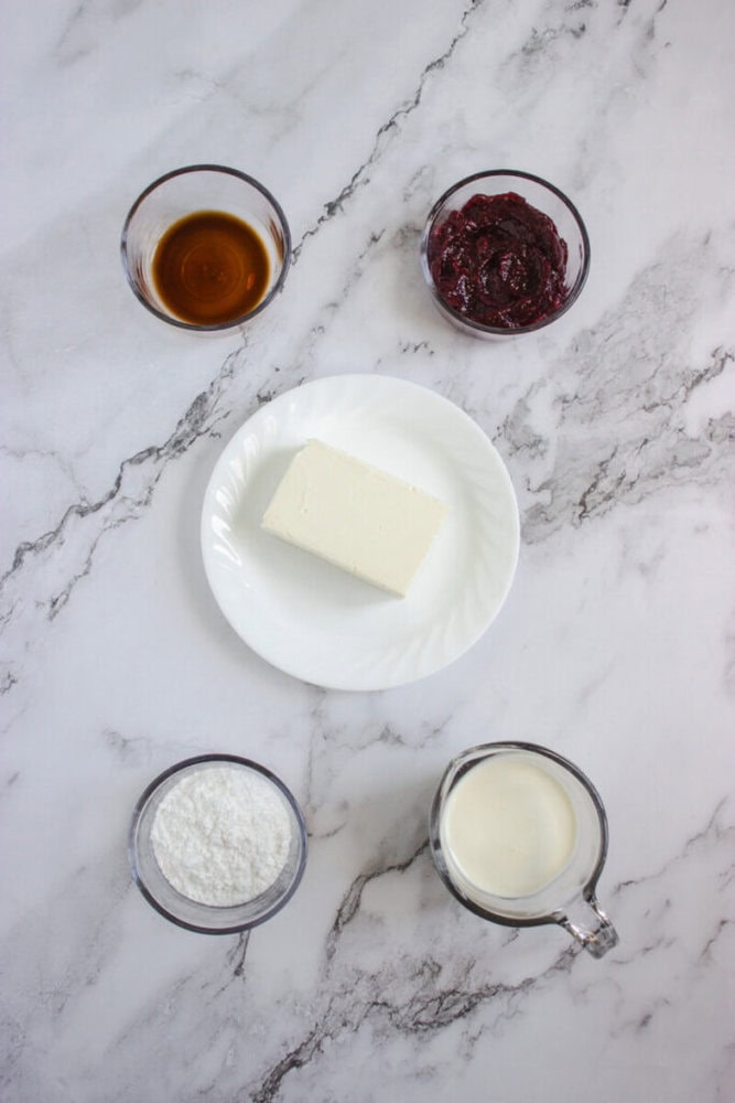 Five ingredients are arranged on a marble surface: a block of cream cheese on a plate, and small bowls of vanilla extract, berry jam, powdered sugar, and milk.