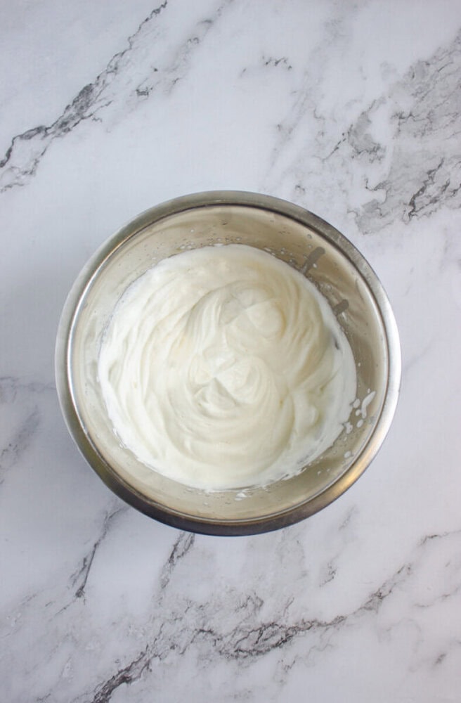 A stainless steel bowl containing whipped cream sits on a white marble countertop.