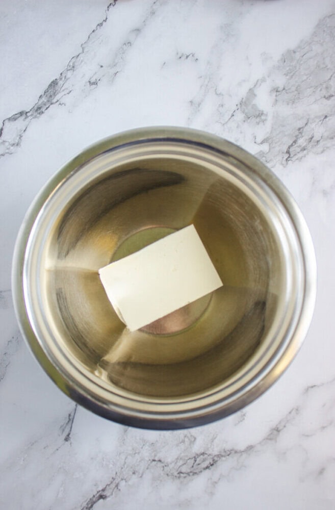 A stick of butter placed in the center of a stainless steel mixing bowl on a marble countertop.