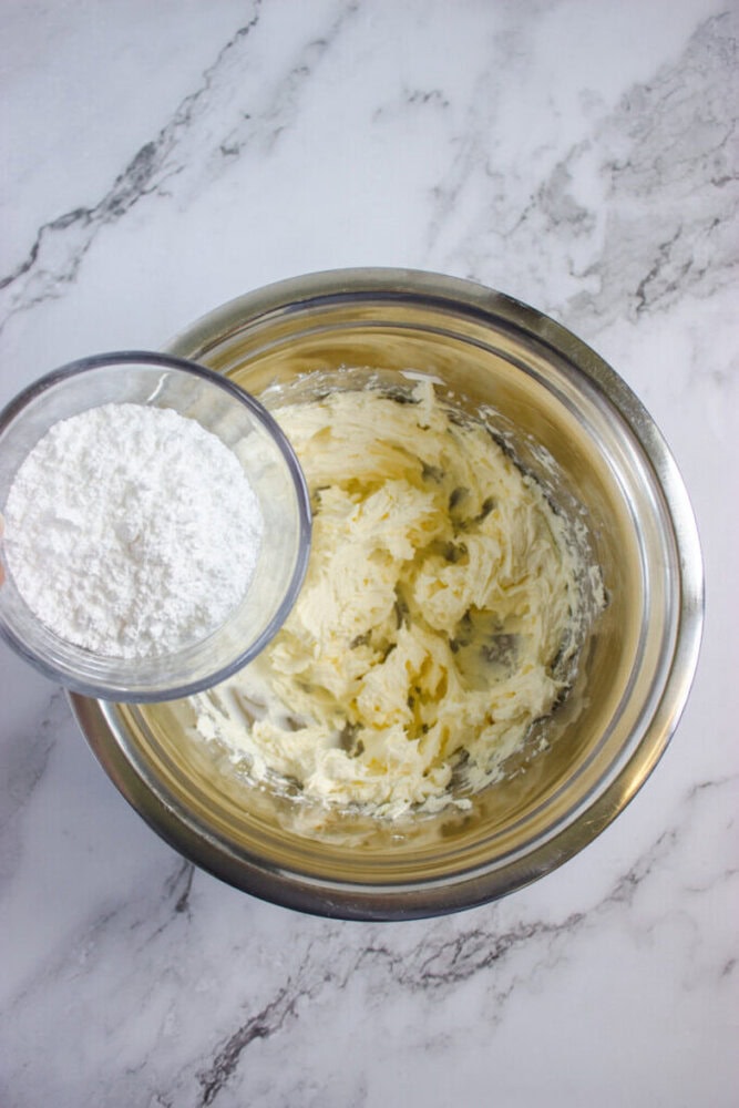 A bowl of creamed butter and sugar with a smaller bowl of powdered sugar being held above it, ready to be added, on a marble surface.