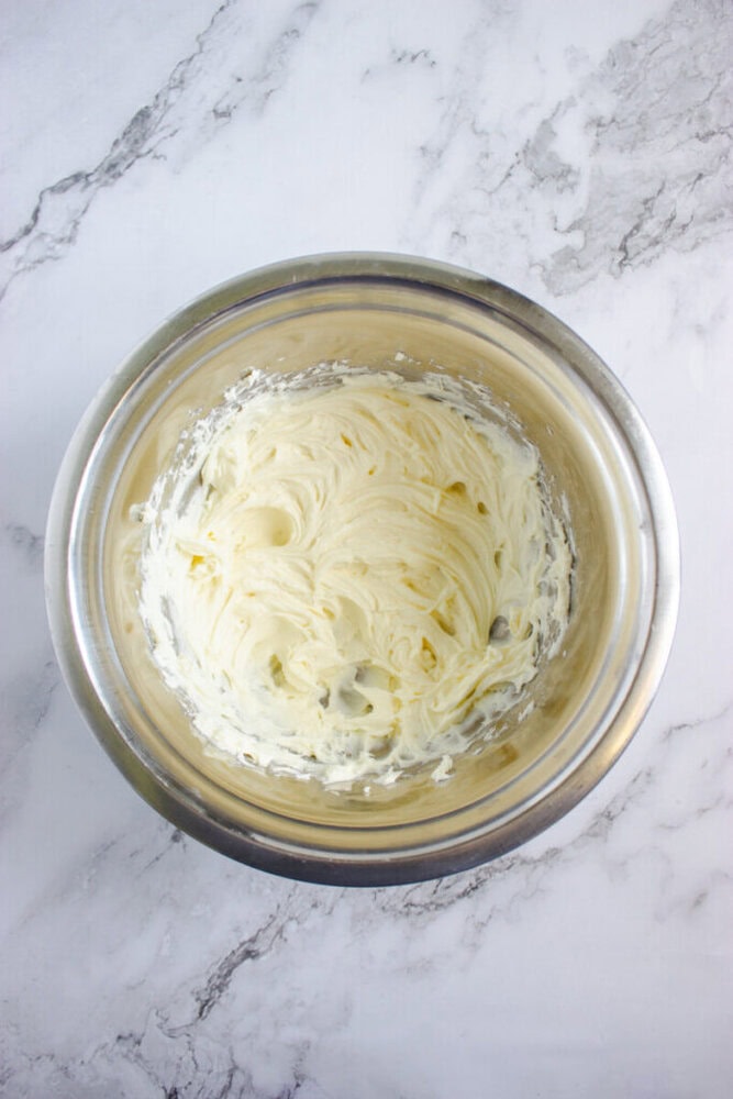 A stainless steel bowl containing whipped cream cheese mixture sits on a marble countertop.