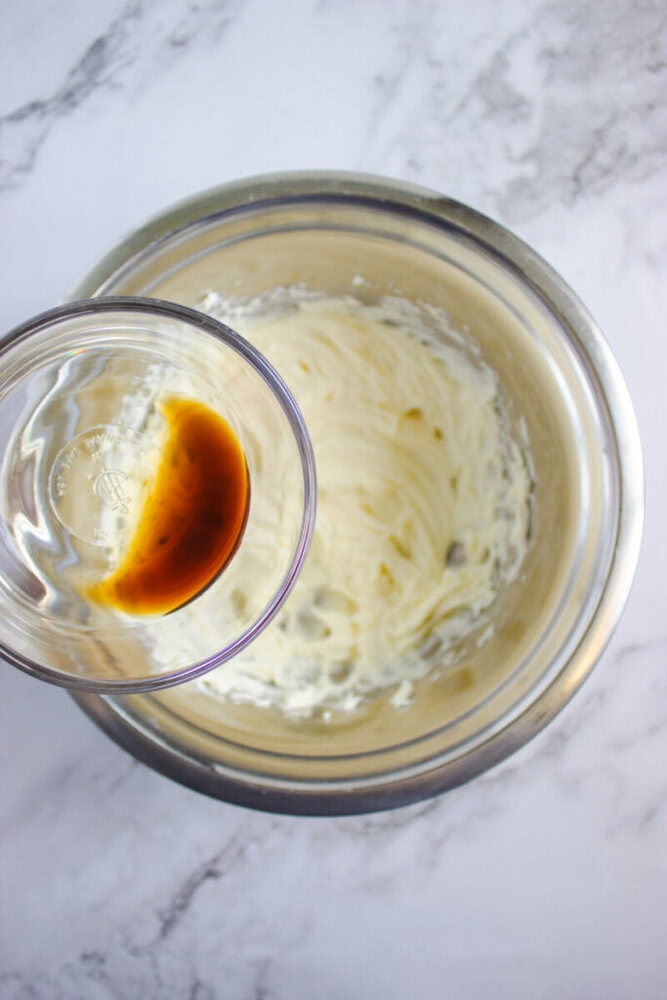 A glass bowl of vanilla extract held above a metal mixing bowl containing creamed butter and sugar on a marble surface.