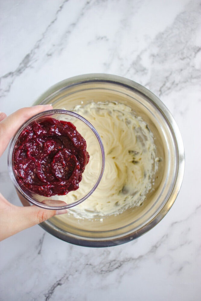 A hand holds a bowl of berry puree above a metal mixing bowl filled with whipped cream, on a marble countertop.
