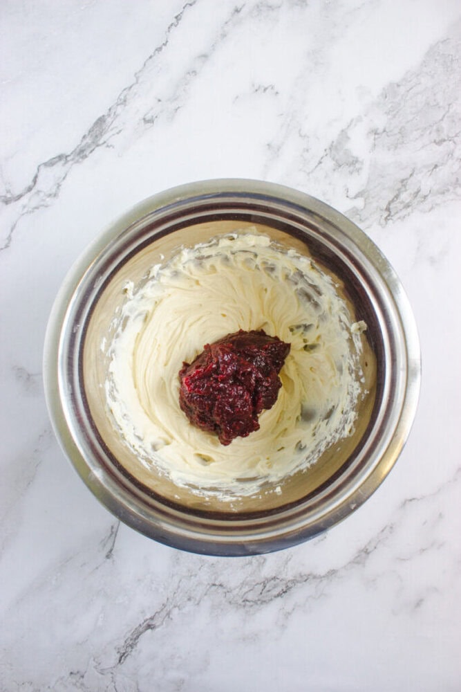 A metal mixing bowl containing creamed butter and sugar with a dollop of berry puree on top, placed on a white marble surface.