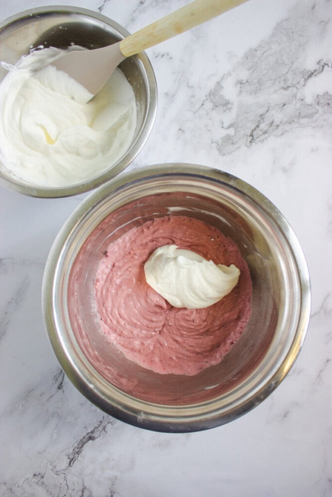 Two metal mixing bowls on a marble surface; one contains whipped cream, the other contains a pink batter with a dollop of whipped cream on top.