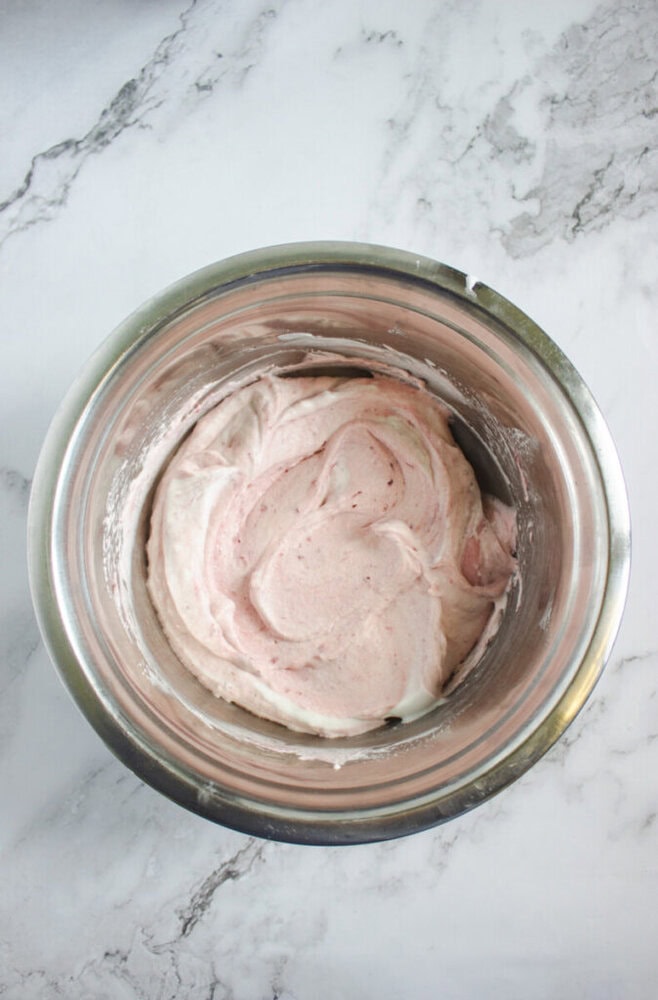 A stainless steel mixing bowl containing a light pink whipped mixture sits on a white marble surface.
