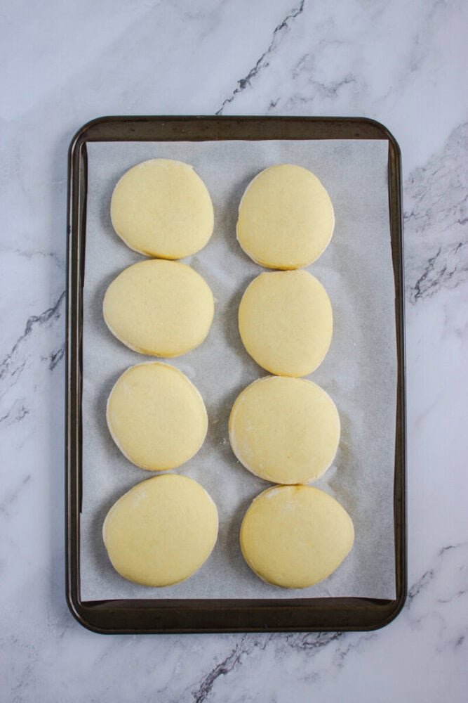 Eight unbaked round dough pieces are arranged in two columns on a parchment-lined baking sheet, set on a marble surface.