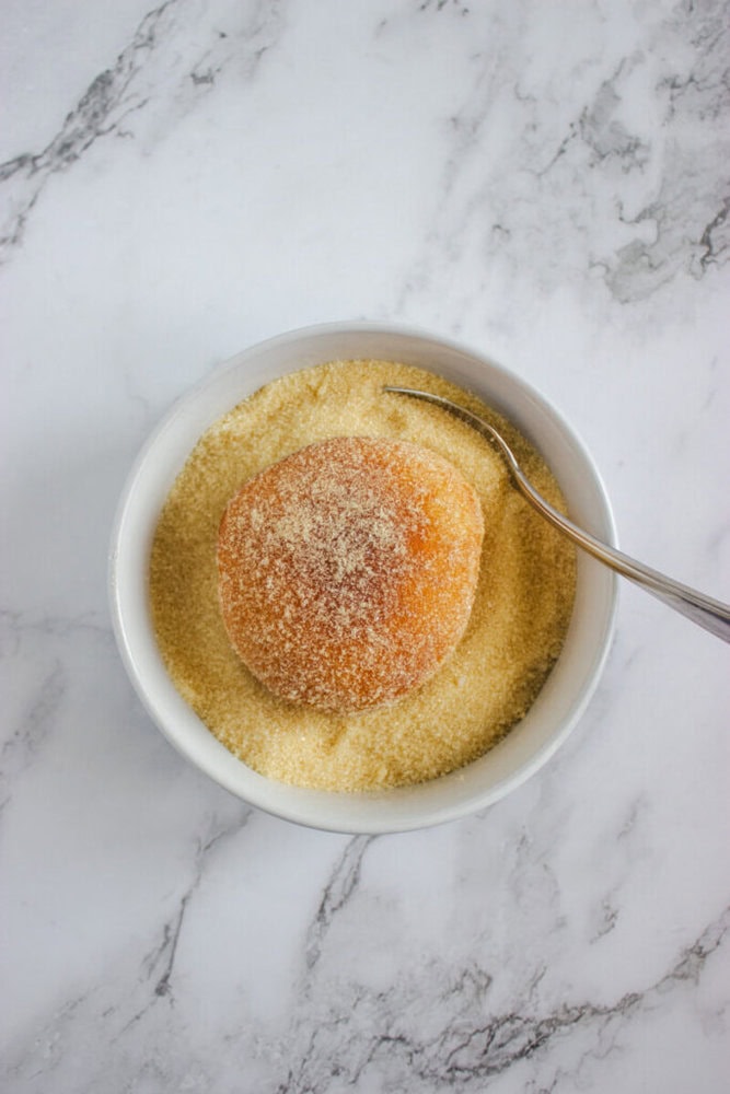 A round donut sits in a white bowl filled with granulated sugar, with a metal spoon resting on the side of the bowl, on a marble surface.