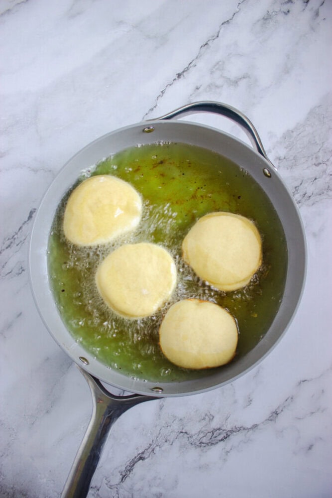 Four round pieces of dough are being deep-fried in hot oil in a frying pan on a marble countertop.