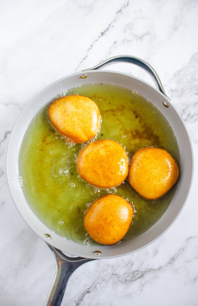 Four round pieces of dough are being deep-fried in hot oil in a gray frying pan on a marble surface.