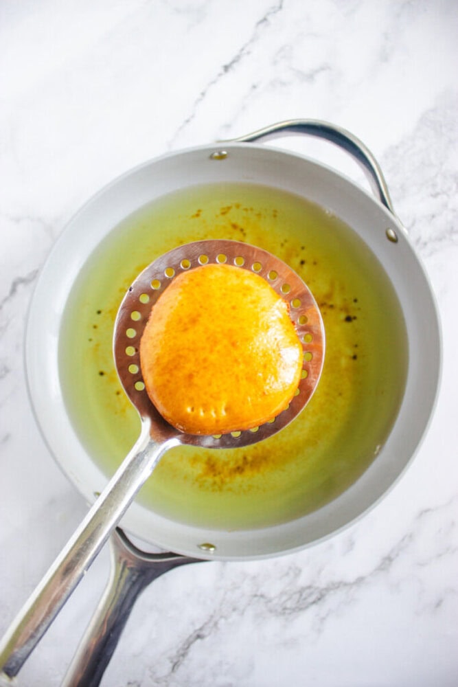 A slotted spoon holds a golden-brown, fried pastry above a pan of hot oil on a marble countertop.