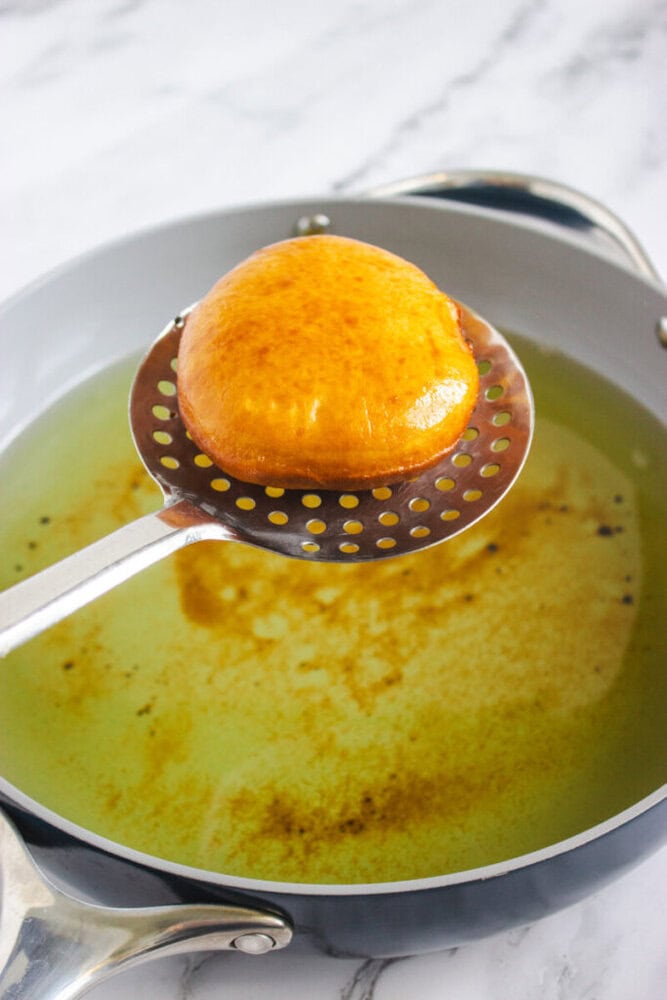 A golden-brown fried dough is held above a pan of hot oil with a slotted spatula.