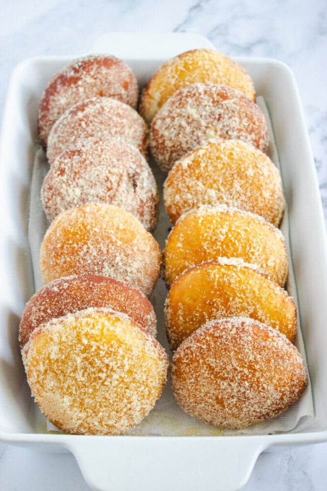 A white rectangular dish filled with rows of sugar-coated round donuts, arranged neatly on a sheet of parchment paper.