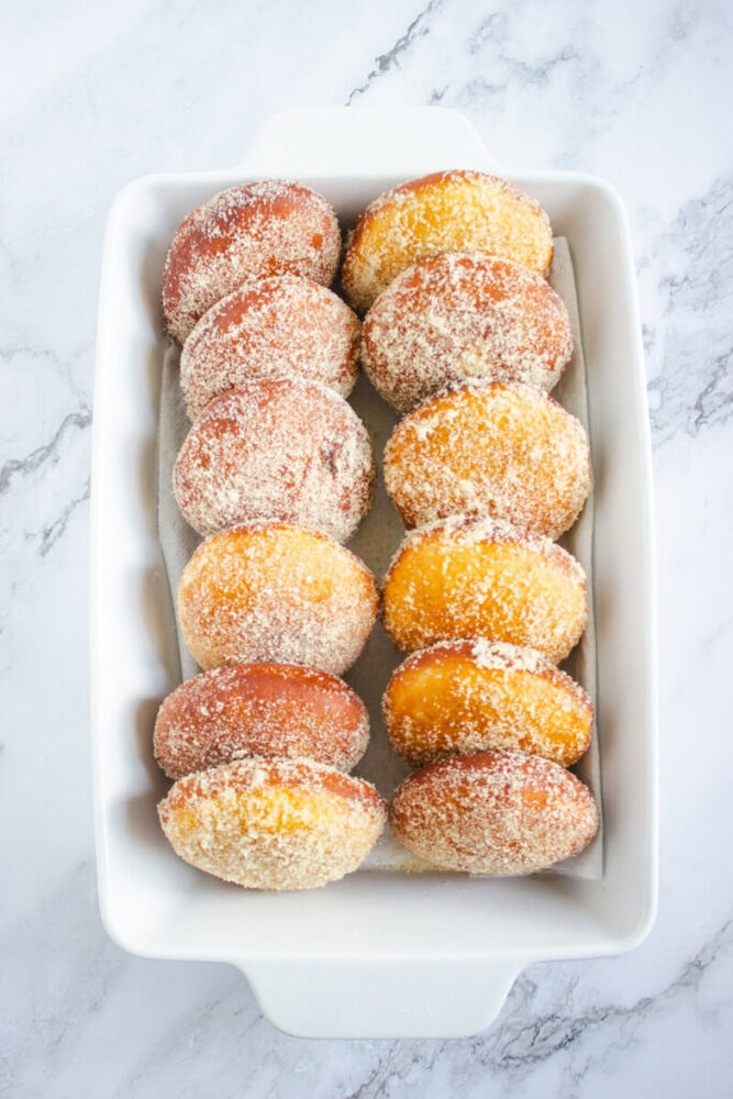 A white rectangular dish containing ten sugar-coated round donuts arranged in two rows on a marble surface.