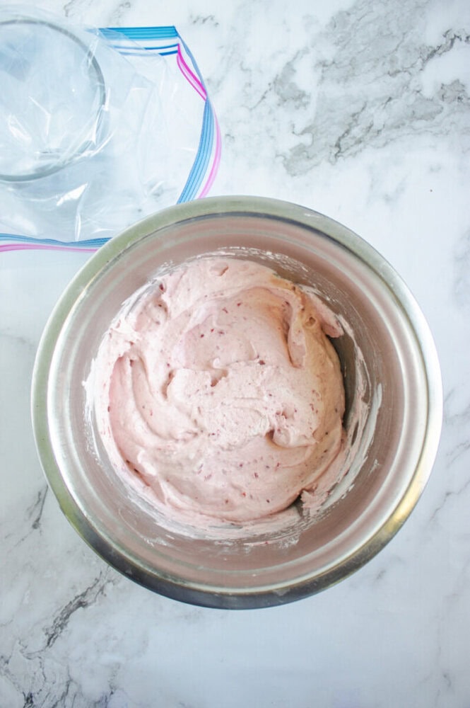 A stainless steel bowl filled with light pink ice cream mixture sits on a marble countertop next to a resealable plastic bag.