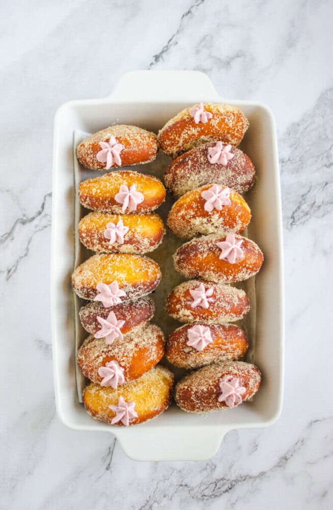 A white rectangular dish holds twelve sugar-coated doughnuts, each topped with a pink flower-shaped icing decoration, on a marble surface.