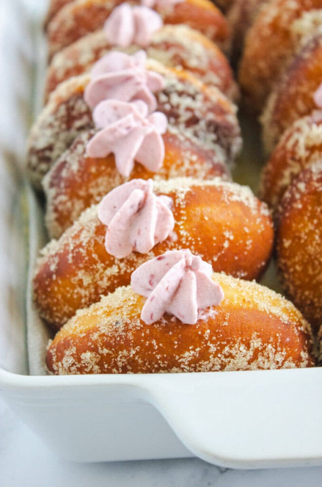 A row of sugar-coated doughnuts topped with pink whipped cream filling arranged in a white dish.