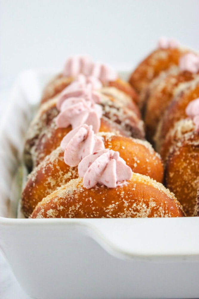 A row of sugar-coated doughnuts in a white dish, each topped with a swirl of pink cream filling.