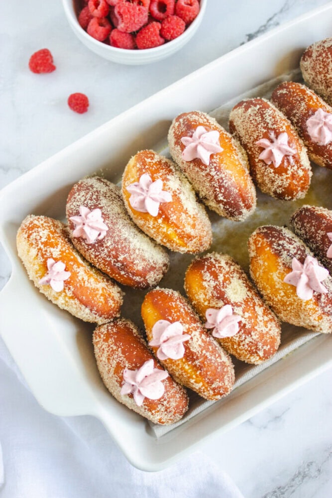 A white baking dish filled with sugared donuts topped with pink cream, with a bowl of raspberries in the background on a marble surface.