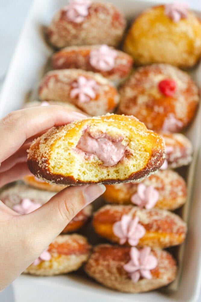A hand holding a bitten doughnut filled with pink cream, with more sugar-coated, cream-filled doughnuts in a tray in the background.