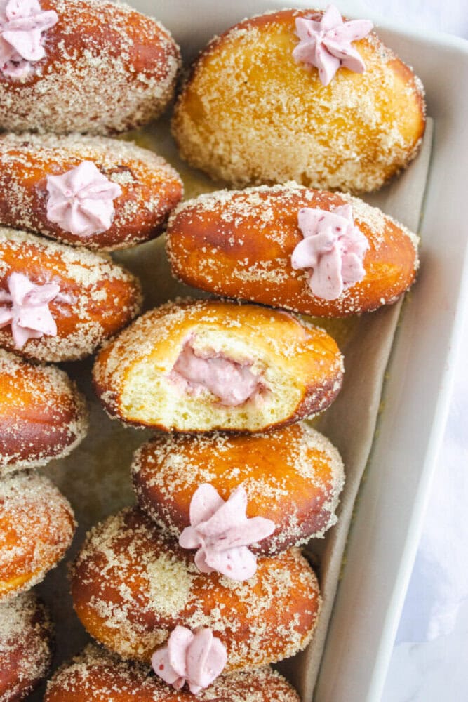A tray of round, sugar-coated donuts, each topped and filled with pink cream. One donut is cut open, showing the creamy filling inside.