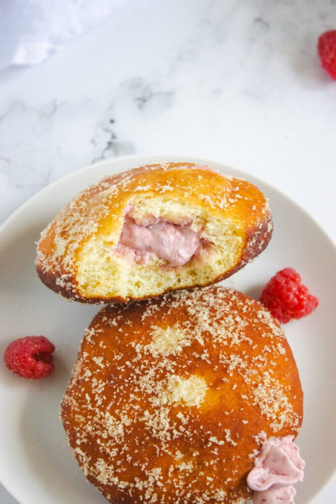 Two sugar-coated donuts on a white plate, one cut open to reveal a pink raspberry cream filling, with whole raspberries on the side.