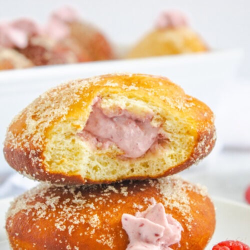 A sugar-coated donut cut in half reveals a pink raspberry cream filling, with whole raspberries and more donuts visible in the background.