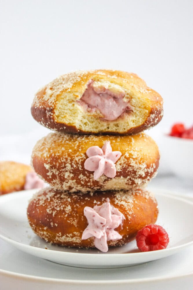 Three sugar-dusted donuts filled with pink raspberry cream are stacked on a white plate, with a fresh raspberry beside them.