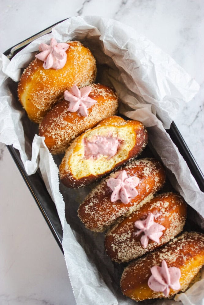 A tray lined with parchment paper holds six sugar-coated doughnuts, each topped with pink cream; one doughnut is cut open, showing pink filling inside.