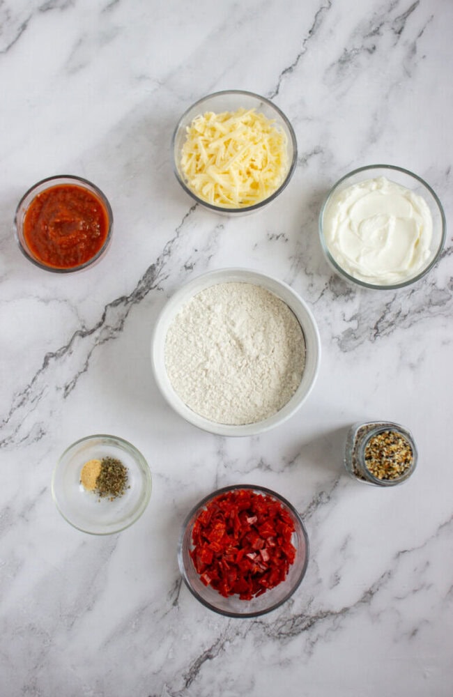 Six glass bowls on a marble surface containing shredded cheese, sour cream, flour, tomato sauce, diced red peppers, and mixed seasonings.