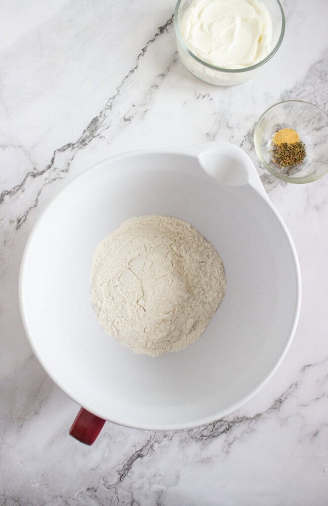 A large white mixing bowl with dry flour mixture on a marble surface, next to a glass bowl of yogurt and a small bowl of herbs and spices.