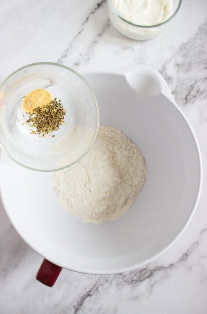 A glass bowl with dried herbs and garlic powder is held above a large white bowl containing flour on a marble countertop.
